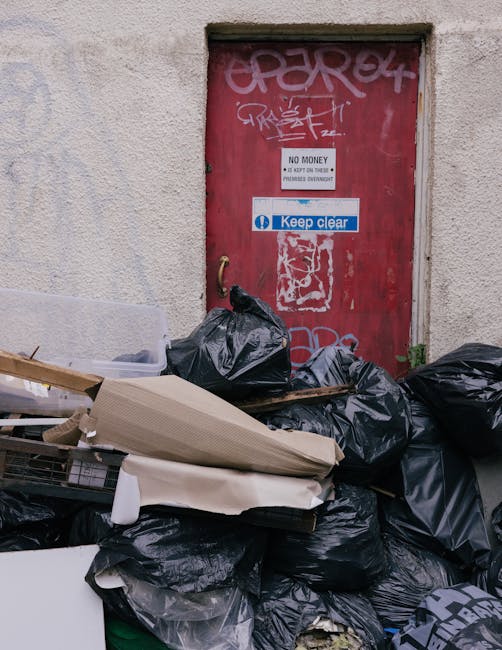 A red metal door with graffiti tags at the top and a 'Keep Clear' sign in the center, set into a beige textured wall. Piled in front of the door are black garbage bags, some torn open revealing their contents, along with discarded cardboard boxes, a partially unrolled paper or plastic sheet, and miscellaneous waste. The area appears cluttered and unclean, with no visible cleaning tools or surfaces, emphasizing the importance of regular waste management and surface cleaning. This image underscores the need for thorough domestic or commercial cleaning to maintain hygiene and tidiness, as promoted by Kentish Town Carpet Cleaning, especially in areas prone to accumulation of waste such as alleyways or building back entrances.