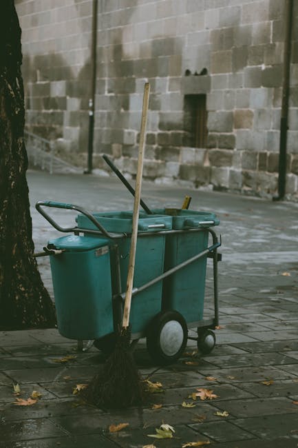 A cleaning trolley with two large turquoise plastic bins, a mop propped against it, and a broom leaning on the side, situated on a paved outdoor surface near a tree with fallen leaves. The background features a beige brick wall with some minor imperfections and small dark spots. The trolley is equipped with wheels for mobility, and cleaning tools are visible, suggesting a deep cleaning or general maintenance setup. This scene is captured in natural daylight, emphasizing the utilitarian nature of the cleaning equipment, which may be used by Kentish Town Carpet Cleaning for surface cleaning and general hygiene maintenance outside residential or commercial properties in Kentish Town.