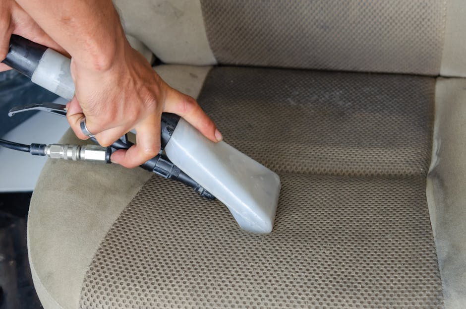 A person is using a handheld upholstery cleaning machine to deep clean a beige fabric car seat in a vehicle interior. The machine's nozzle is pressed onto the seat surface, which features a textured pattern with a mix of light and dark beige tones, indicating a thorough surface cleaning process. The surrounding area shows the seat's upholstery, with visible dirt and stains, highlighting the importance of professional cleaning for hygiene and maintenance. The scene is well-lit, emphasizing the equipment and the fabric's condition, with a focus on surface cleaning and sanitisation carried out by Kentish Town Carpet Cleaning, reflecting their expertise in domestic cleaning services for upholstery in Camden Council-permitted environments.