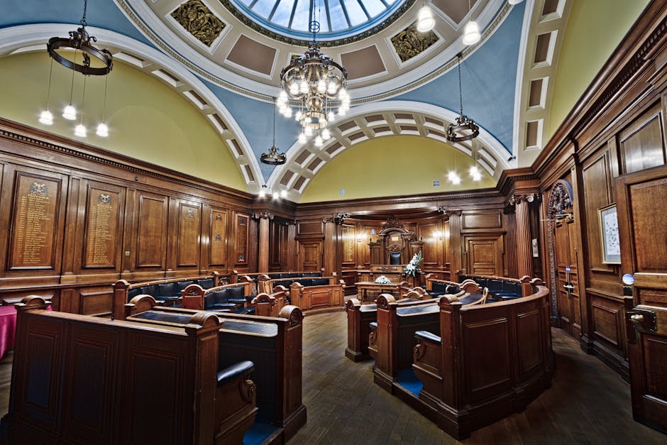 Interior view of a historic council chamber with polished wooden paneling on the walls, arranged in a semi-circular layout with black leather-upholstered seats and wooden desks. The room features a high, domed ceiling painted in pastel shades of yellow, blue, and white, with ornate decorative molding and a large central skylight allowing natural light to illuminate the space. A multi-tiered chandelier hangs from the ceiling, providing additional lighting. The overall appearance of the room is clean, well-maintained, and showcases detailed woodwork, reflecting a professional approach to surface cleaning and heritage room preservation by Kentish Town Carpet Cleaning, aligning with the theme of surface cleaning and hygiene for Camden Council permits.