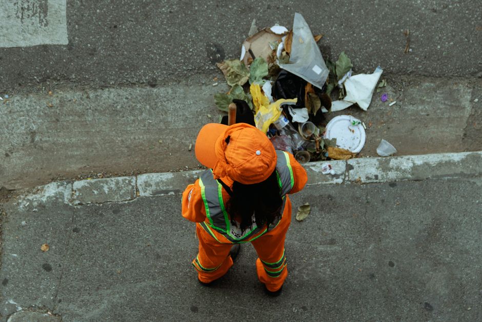 A sanitation worker dressed in bright orange high-visibility clothing and a matching hat, viewed from above, standing on a concrete pavement beside a curb. The worker is facing a pile of mixed waste, including paper, plastic, and organic debris, gathered against a concrete wall. The scene is outdoors and illuminated by natural light, with the worker appearing to assess or prepare to clean up the waste. The image relates to surface cleaning and waste management, emphasizing hygiene and sanitation practices, relevant to Camden Council rules for waste disposal from cleaning jobs, as featured on kentishtowncarpetcleaning.com, where Kentish Town Carpet Cleaning provides domestic and commercial cleaning services.