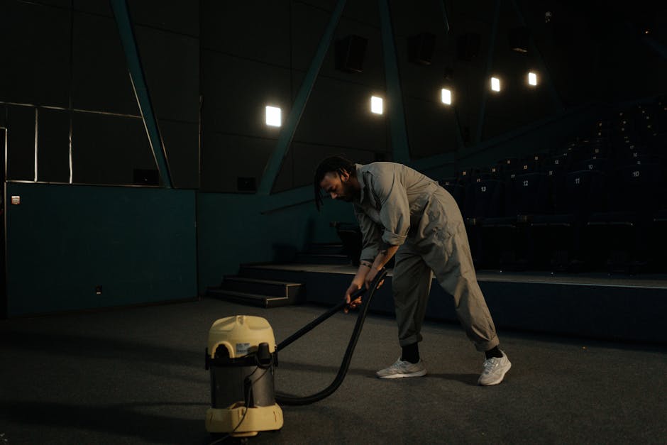A man wearing a light grey jumpsuit and white sneakers is using a yellow and black industrial vacuum cleaner to clean a dark carpeted floor in a dimly lit auditorium or conference room. He is standing slightly bent forward, holding the vacuum hose with both hands, with the vacuum placed on the ground in front of him. The room features dark blue tiered seating with numbered markers, a high ceiling with industrial lighting fixtures, and a backdrop of dark walls and stairs. The scene emphasizes surface cleaning and deep cleaning processes, with a focus on maintaining hygiene and cleanliness in large indoor spaces, as demonstrated by Kentish Town Carpet Cleaning.