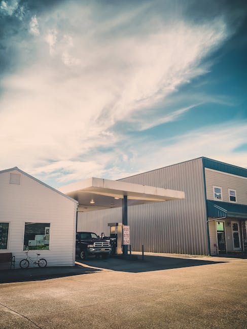 A commercial building with grey metal siding and white trim, featuring multiple windows and a small attached office area, located in a parking lot with a covered fueling station. A black pickup truck is parked underneath the canopy, and a bicycle is seen leaning against the white building. Bright, natural daylight illuminates the scene, with a partly cloudy sky overhead. The image exemplifies clean, maintained exterior surfaces typical of a commercial property, reflecting the high standards of professional cleaning services provided by Kentish Town Carpet Cleaning, reinforcing the focus on surface cleaning and building sanitisation near Kentish Town station with same day service.