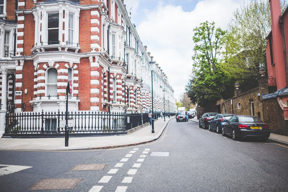 A street view of Victorian-style terraced buildings with intricate white and red brick facades, large bay windows, and decorative architectural details along Castlehaven Road. The sidewalk is clean and well-maintained, with a black wrought-iron fence enclosing a small garden area. Several parked cars line the curb, including sedans and hatchbacks, with some reflecting sunlight on their polished surfaces. The street is empty of pedestrians, and the day appears partly cloudy with some blue sky visible above lush green trees lining the sidewalk. The image captures a quiet, residential area in NW5, KENTISH TOWN, showcasing well-preserved historic architecture and a tidy urban environment, typical of the surroundings where Kentish Town Carpet Cleaning provides expert surface cleaning, deep cleaning, and sanitation services.
