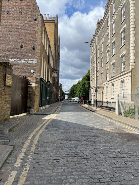 A cobblestone street in NW5, London, lined with historic brick and stone buildings on both sides. The street appears clean, with two yellow lines along the curb indicating parking restrictions. Overhead, partly cloudy skies cast natural light over the scene. Visible features include a lamppost, parked cars in the distance, and trees providing greenery along the sidewalk. This setting reflects the urban environment typically serviced by Kentish Town Carpet Cleaning for surface cleaning, deep cleaning, and sanitisation of residential or commercial premises in the Castlehaven Road area.