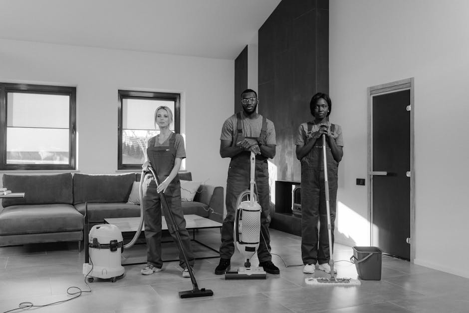 Three professional cleaners from Kentish Town Carpet Cleaning are positioned in a modern living room, engaged in surface cleaning activities. The room features a large L-shaped sofa with fabric upholstery, two windows allowing natural light, and a dark wooden panel wall. The cleaners are dressed in grey uniforms, each holding cleaning tools: one with a vacuum hose, another with a steam mop, and the third with a spray bottle and cloth. The floor is made of polished tiles, appearing clean and reflective, indicating recent deep cleaning or sanitisation. Various cleaning equipment, such as a vacuum cleaner, a mop, and a small bucket, are visible, suggesting thorough domestic cleaning and surface maintenance. The room appears well-lit with soft natural light, highlighting the sleek surfaces and cleanliness achieved by Kentish Town Carpet Cleaning, ensuring hygienic and visually appealing living spaces. This image exemplifies professional cleaning processes aimed at maintaining hygiene and surface care within residential environments.