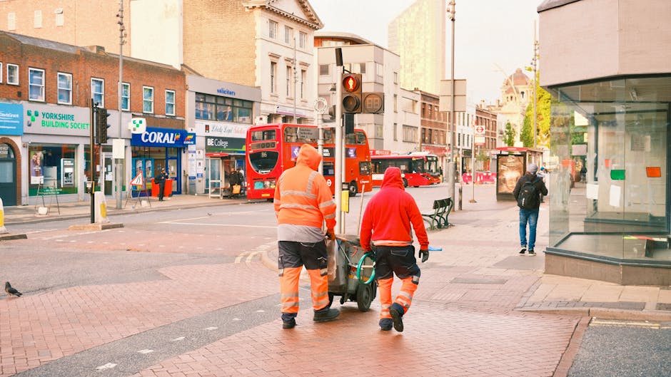 Two street cleaning workers wearing high-visibility orange uniforms with reflective stripes are walking along a busy urban sidewalk in Kentish Town, London, near a bus stop and a pedestrian crossing. One worker is pushing a small cleaning cart, while both are walking in front of a traffic light showing red. The scene includes a double-decker bus, various storefronts, and pedestrians. The bright morning or late afternoon lighting highlights the cleanliness and orderliness of the area, with the pavement and surrounding surfaces appearing clean and well-maintained. Kentish Town Carpet Cleaning emphasizes surface cleaning and sanitisation services for local residential and commercial spaces, ensuring hygienic environments.