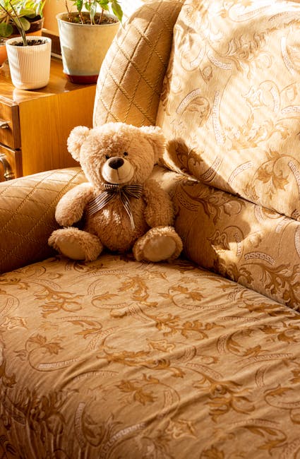 A close-up of a beige patterned fabric sofa in a living room, featuring intricate floral and scroll designs on the upholstery. Resting on the sofa is a plush teddy bear with light brown fur, a dark ribbon tied around its neck, and a friendly facial expression. The background shows a wooden side table with several potted plants, including a large cactus in a light-colored pot, and soft natural light illuminating the scene. The setting appears clean and well-maintained, with a focus on home comfort and domestic surface cleaning, aligning with the services offered by Kentish Town Carpet Cleaning for upholstery deep cleaning and sanitisation.