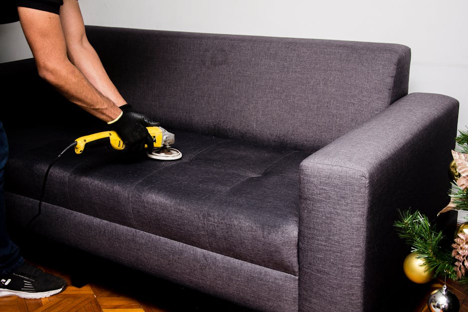 A person wearing black gloves is using a handheld electric scrubber to deep clean and refresh the dark grey upholstered fabric of a sofa in a living room. The sofa has a simple, modern design with armrests on both sides and is situated on a polished wooden floor. The background shows a plain, light-colored wall, and part of a decorated Christmas tree with gold and pink ornaments is visible to the right. The scene emphasizes surface cleaning and upholstery sanitisation, highlighting the meticulous cleaning process undertaken by Kentish Town Carpet Cleaning as part of their domestic cleaning services.