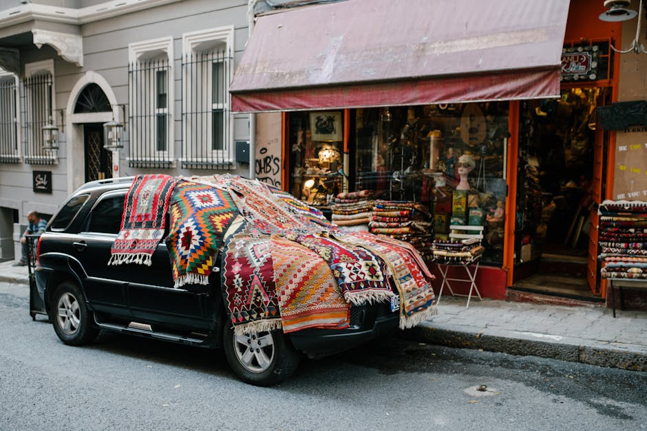 A black SUV parked on a city street with its hood covered by numerous colorful, patterned rugs and textiles, partially draping over the front and sides. The vehicle is positioned in front of a shop with a red awning and large glass windows displaying various items, including stacked fabric or clothing. The surrounding area features classic European-style buildings with white facades and black wrought-iron balconies. The street appears clean and quiet, with a pedestrian visible in the background. This scene highlights detail and smoothness in the textiles, with natural daylight illuminating the scene, and the fabrics exhibiting vibrant colors and intricate patterns, suggestive of upholstery or decorative rugs associated with domestic or commercial interior decorating. Kentish Town Carpet Cleaning offers surface cleaning and deep cleaning services, focusing on maintaining hygienic and pristine environments.