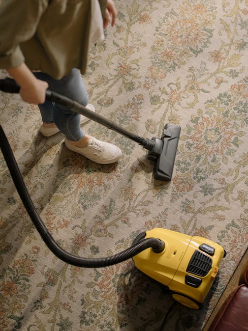 A person operating a yellow and black vacuum cleaner with a long hose attachment on a patterned beige and green area rug in a room. The individual is wearing grey jeans, beige sneakers, and a tan jacket, and is guiding the cleaning device across the carpet. The room features wooden flooring visible around the rug, and the lighting is soft and natural, highlighting the clean and well-maintained appearance of the carpet. This scene illustrates domestic surface cleaning, which is part of comprehensive sanitisation and deep cleaning services offered by Kentish Town Carpet Cleaning to ensure hygiene and maintenance of residential spaces.