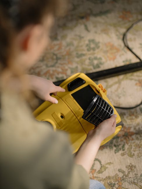 A person operating a yellow portable carpet cleaning machine with a black vent cover, positioned on a patterned, beige and green rug in a room with wooden flooring. The individual, seen from behind, holds the device with both hands, with a focused posture. The room is well-lit, highlighting the clean, maintained surfaces, and there are visible wires connected to the cleaning equipment. This scene exemplifies deep surface cleaning and sanitisation services offered by Kentish Town Carpet Cleaning, emphasizing their prompt and professional domestic cleaning solutions.