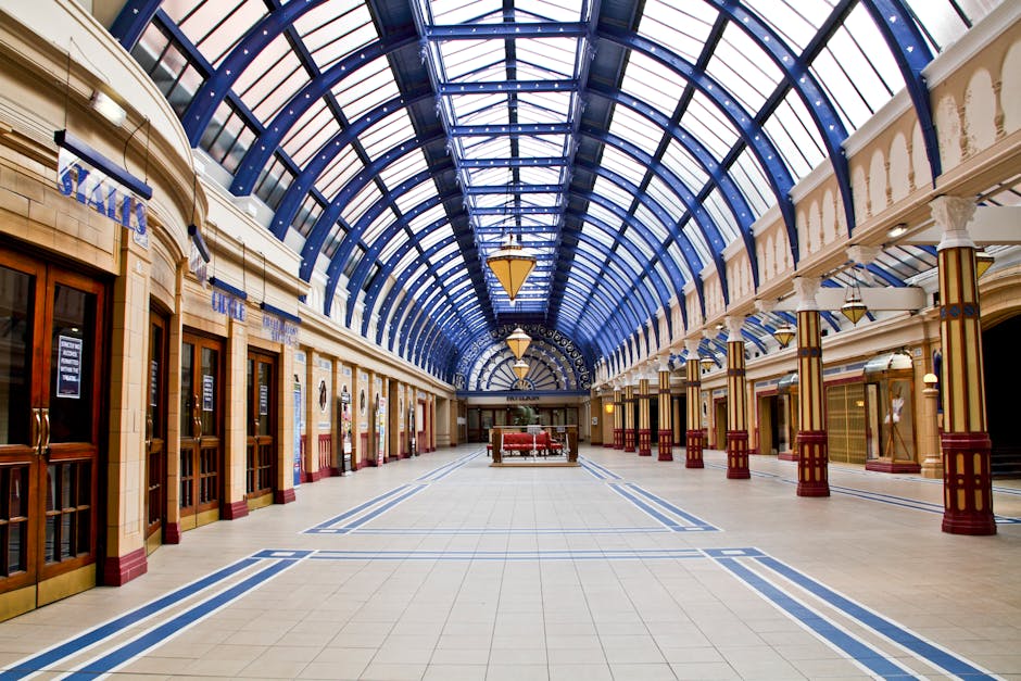 Interior view of the Victorian-style shopping arcade featuring a large, arched glass ceiling with blue framing, allowing natural light to illuminate the tiled floor. The wide corridor is lined with wooden storefronts, each with glass doors and signage, and decorative columns with gold accents supporting the structure. The mall appears well-maintained, with clean and polished floors, and the space is evenly lit by both natural and ambient lighting, creating a bright and inviting atmosphere. The perspective showcases the symmetry of the architecture, emphasizing the spaciousness and ornate design elements typical of the historic building, which Kentish Town Carpet Cleaning maintains through regular deep cleaning and surface sanitisation services.