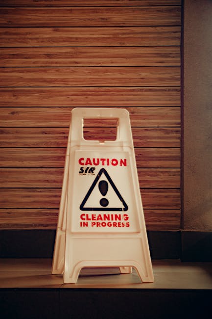 A white plastic 'Caution Cleaning in Progress' floor sign featuring a black hazard triangle with an exclamation mark in the center, placed on a smooth wooden floor next to a textured wall with horizontal wood paneling. The sign is positioned at the edge of the room, indicating ongoing cleaning activity. The lighting is moderate, highlighting the cleanliness of the area, which appears well-maintained. This image, associated with Kentish Town Carpet Cleaning, emphasizes the importance of surface cleaning and hygiene during end of tenancy cleaning in NW5, KENTISH TOWN.