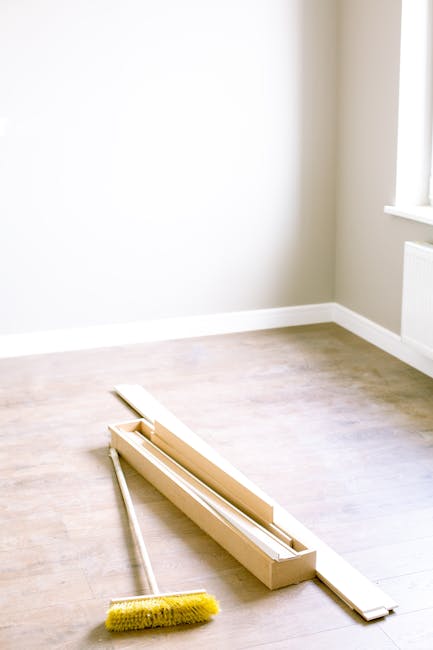 A bright, empty room with light-colored wooden flooring and a white wall with a window allowing natural light. On the floor, there is a wooden window frame with a sliding mechanism, positioned near the corner of the room. A yellow cleaning brush with a long handle is placed next to the window frame, suggesting a recent or ongoing cleaning process. The room appears clean and well-maintained, with no visible dust or clutter. This scene showcases typical surface cleaning and preparation for further deep cleaning or maintenance by Kentish Town Carpet Cleaning, emphasizing the importance of hygiene in domestic cleaning routines.